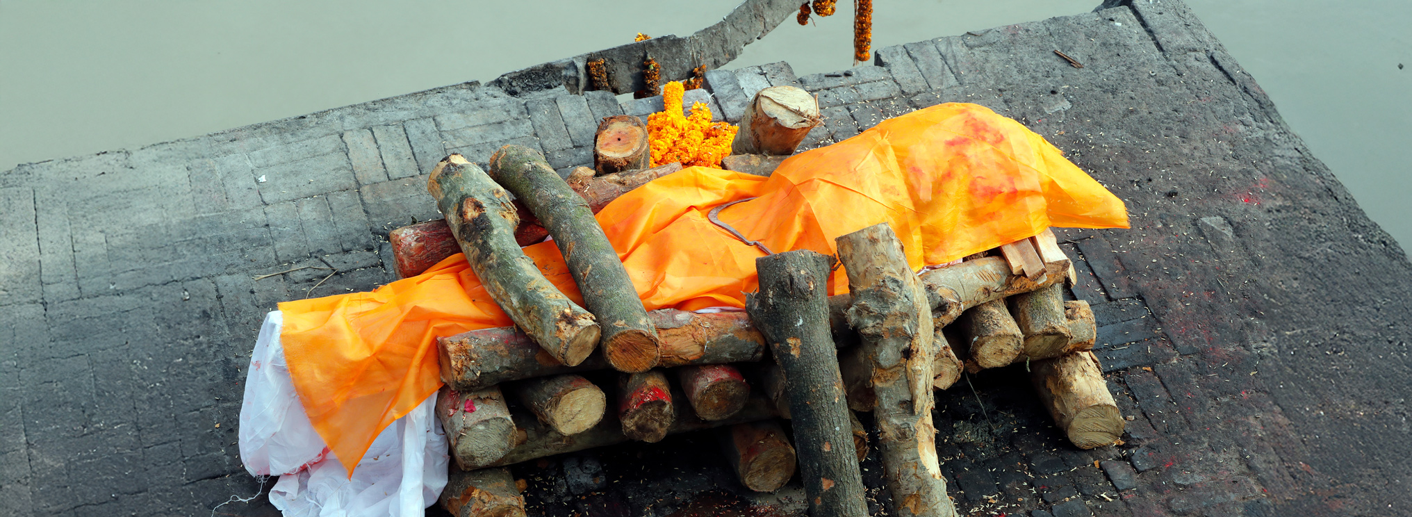 Ein mit einem orangefarbenen Tuch bedeckter Körper ruht auf einer Holzplattform für das rituelle Verbrennungsritual. Die Szene zeigt eine traditionelle hinduistische Feuerbestattung als Teil des spirituellen Abschieds und der Vorstellung von Wiedergeburt.